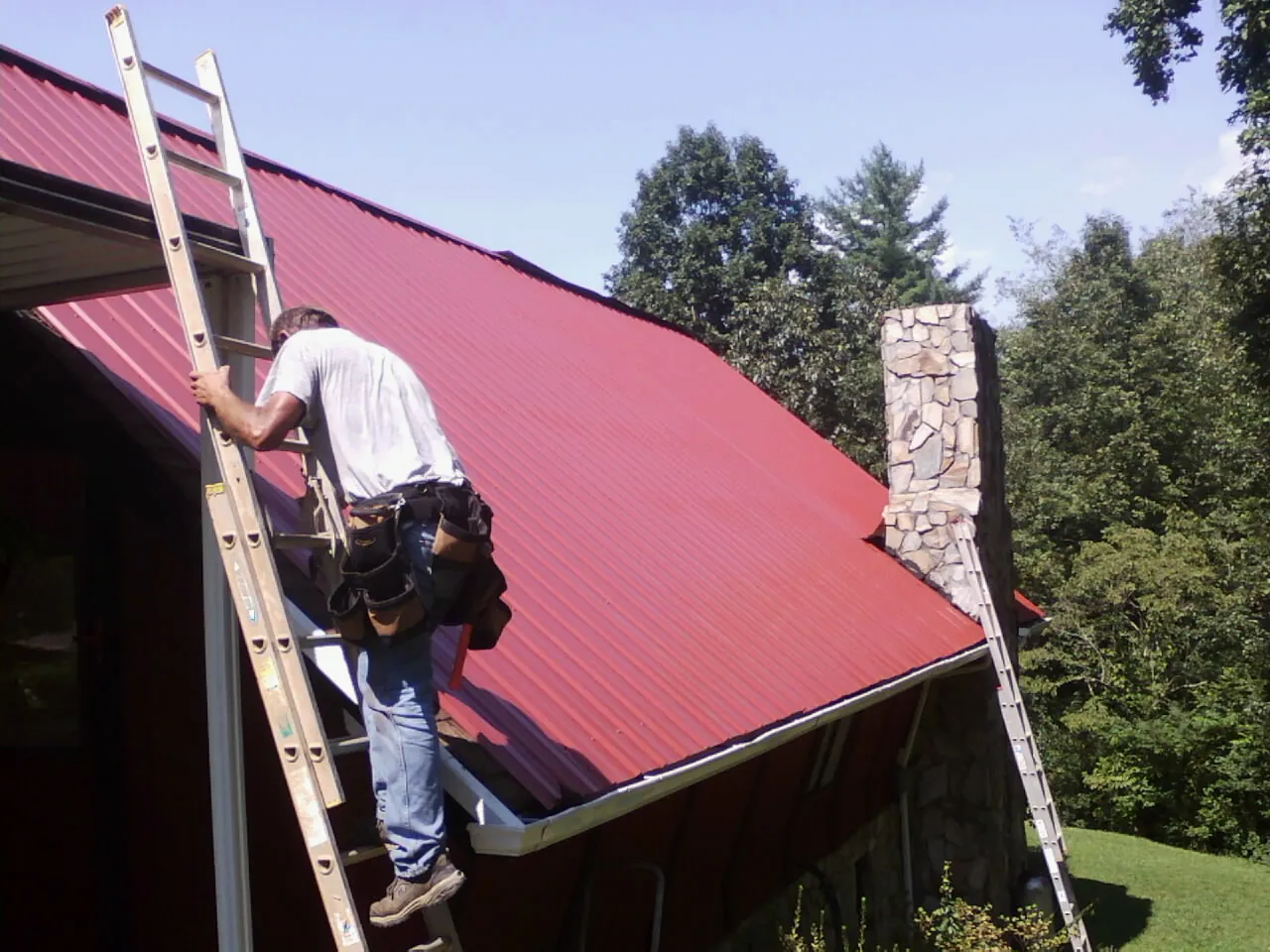 Roofer on a ladder beside a red metal roof and stone chimney — Soffit & Fascia Repair by Roofing Brothers in South Milford
